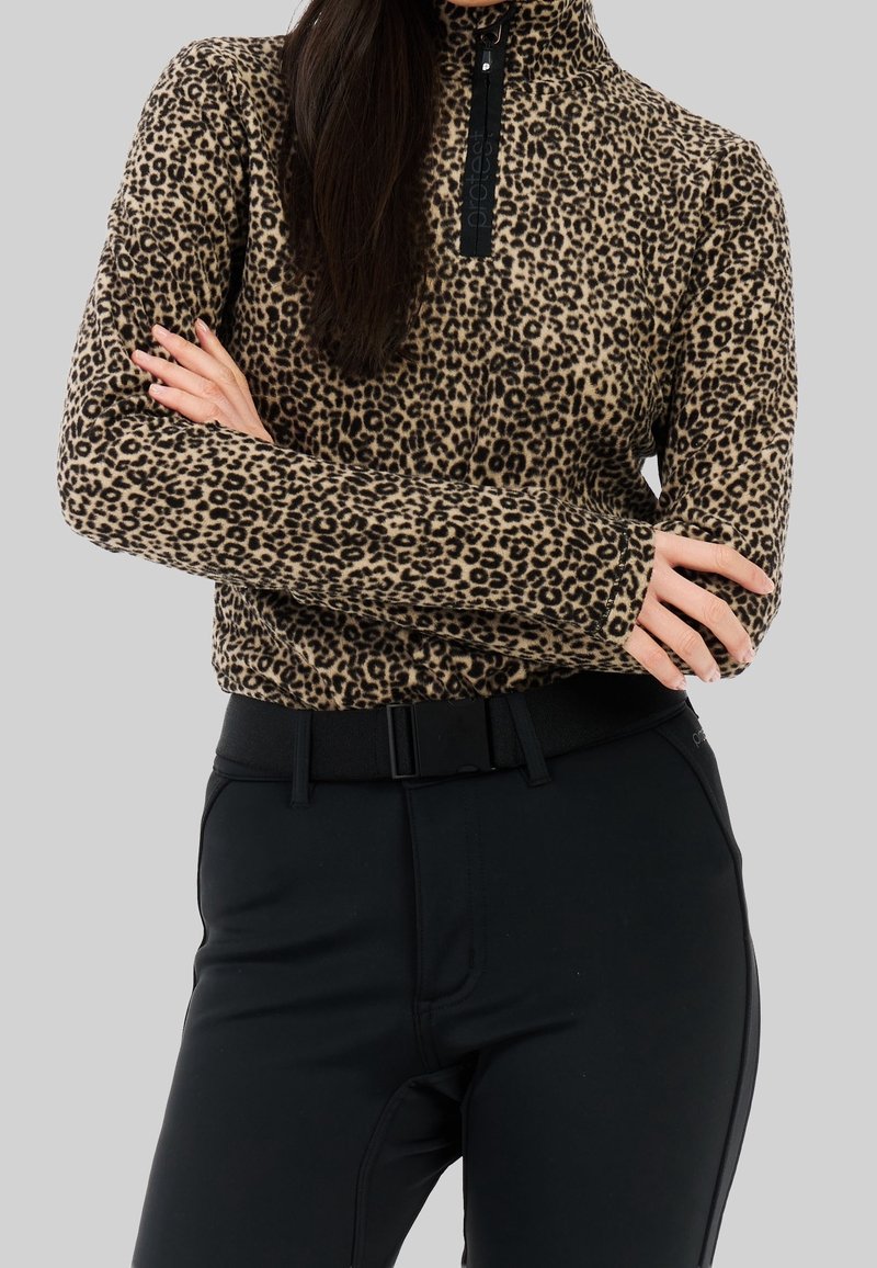 Femme portant un pull à manches longues à imprimé léopard avec une fermeture éclair noire et un pantalon noir, bras croisés devant un fond gris.