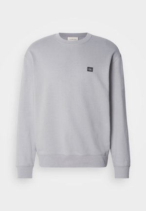 Sudadera gris claro hecha de una mezcla de algodón. Presenta mangas largas, cuello redondo y un pequeño parche con el logo en el pecho. Textura suave.
