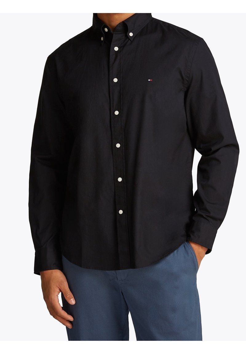 Homme portant une chemise noire boutonnée avec un petit logo sur la poitrine, associée à un pantalon bleu, mains détendues le long du corps.
