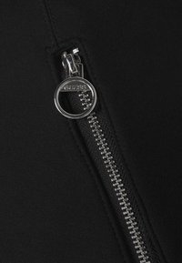 Zwarte stof met een metallic zilveren rits met een ronde trekker met het label "GUESS." Het oppervlak heeft een gladde textuur met zichtbare stiksels.