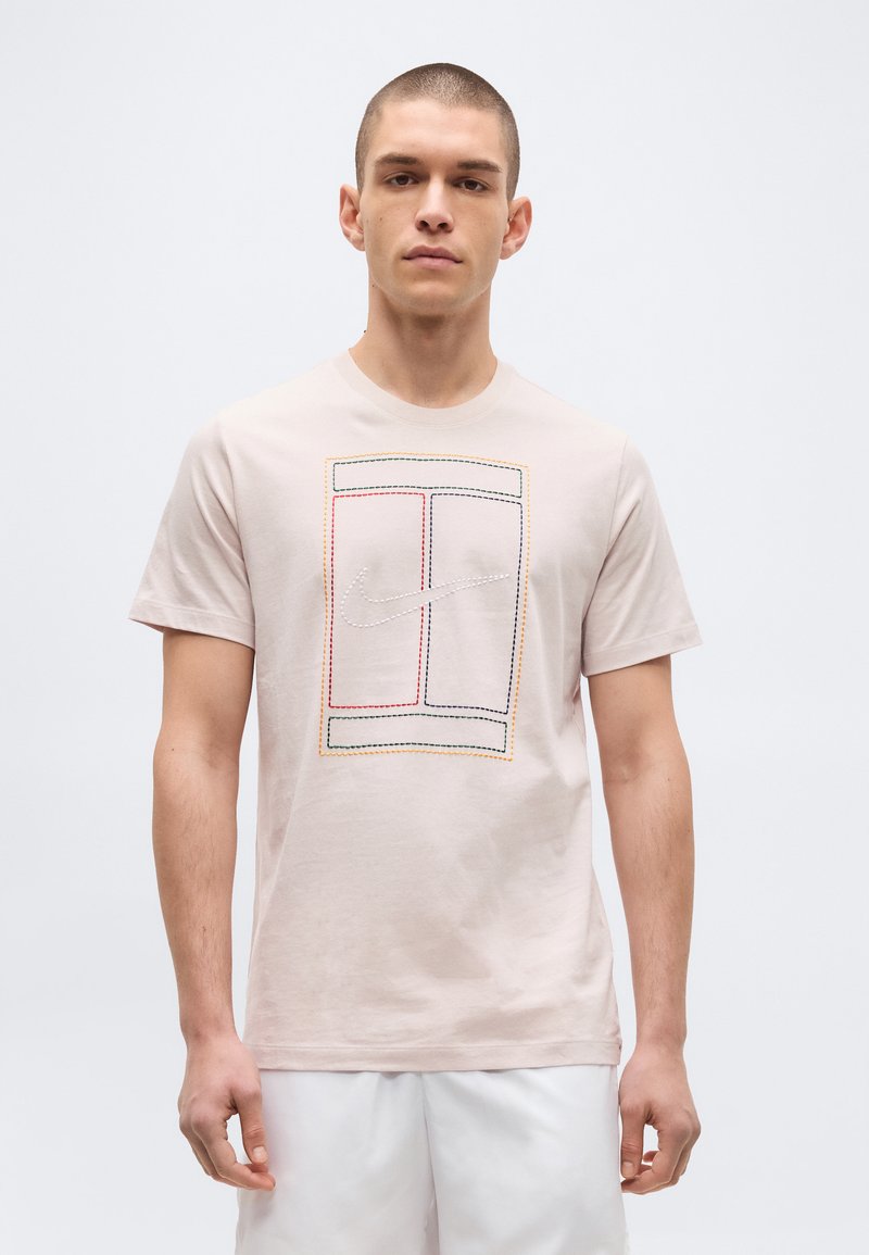Jeune homme portant un t-shirt beige clair avec un design rectangulaire multicolore cousu et un logo Nike swoosh au centre, assorti à un short blanc.