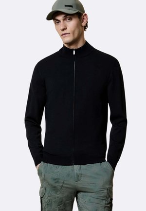 Giovane uomo che indossa un maglione nero con zip, pantaloni cargo verdi con motivo e un berretto grigio, in piedi con le mani in tasca.
