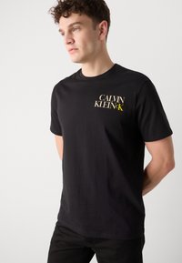 Sort bomuld t-shirt med korte ærmer, der har "CALVIN KLEIN" logo i beige og gul på fronten. Klassisk rund hals design.