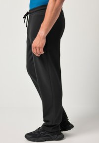 Zwarte joggingsbroek met elastische tailleband en trekkoord. Bevat zijzakken en heeft een taps toelopend ontwerp. G gedragen met zwarte sportschoenen.