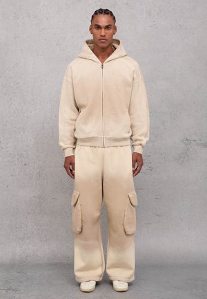 Sudadera con cremallera en color beige, con un bolsillo frontal y pantalones cargo a juego que tienen bolsillos laterales, elaborados con una tela suave y texturada. Zapatillas blancas.