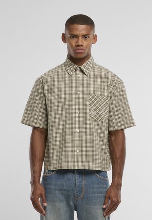 Jeune homme portant une chemise à manches courtes beige et marron à carreaux avec poche poitrine, associée à un jean bleu, debout devant un fond uni.