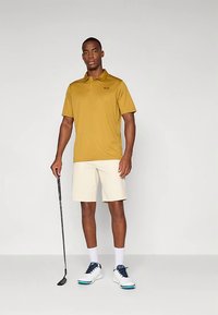 Krátky rukáv horčicovožltá polokošeľa, krémové šortky, čierne golfové palice, biele topánky s modrými akcentmi. Model stojí na neutrálom pozadí.