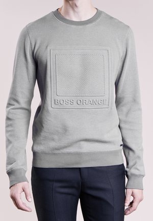 Mann trägt hellgraues, strukturiertes Langarm-Sweatshirt mit geprägt eingeprägtem Quadrat und dem Schriftzug „BOSS ORANGE“, kombiniert mit dunkler Hose.