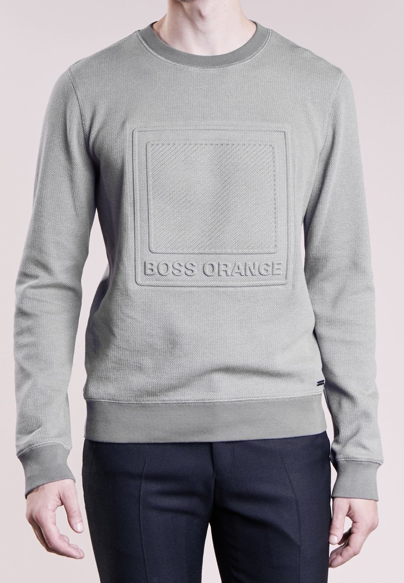 Homme portant un sweat-shirt gris clair texturé à manches longues avec un carré embossé et le texte « BOSS ORANGE », associé à un pantalon foncé.