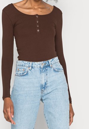 Kvinde iført en brun ribbet langærmet top med knapdetaljer, stukket ned i højttaljede lyseblå denimjeans.