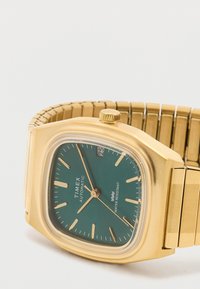 Reloj automático Timex en tono dorado con esfera verde, agujas y marcadores dorados, pantalla de fecha y pulsera metálica expandible dorada.