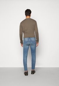 Hombre de pie de espaldas, vistiendo una camisa marrón de manga larga, jeans ajustados azul claro y zapatos de cuero negros sobre un fondo liso.