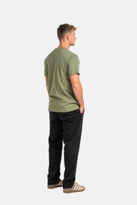 Camiseta de algodón verde oliva de manga corta, combinada con pantalones negros. El modelo está de lado, usando zapatillas marrones con rayas blancas.