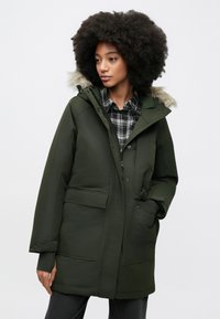 Groene gewatteerde parka met een capuchon gevoerd met nepbont, meerdere zakken en een ritssluiting aan de voorkant. Gedragen over een zwart-wit geruit overhemd.