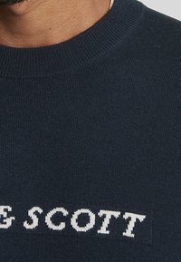 Vista parziale di una persona che indossa un maglione di lana di un blu navy scuro con il testo bianco "E SCOTT" visibile nella zona del petto.