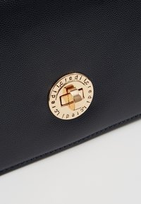 Námořnická kožená kabelka s texturovaným povrchem, kulatým zlatým kováním s uzávěrem a vyraženým názvem značky po obvodu.