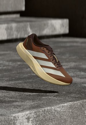 Brązowy i kremowy but do biegania Adidas z białymi paskami unoszący się nad szarymi betonowymi schodami na zewnątrz.
