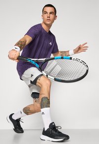 Lilla tekstureret sports T-shirt, hvidt armbånd, sorte shorts og sorte tennissko. Holder en tennisracket med blå detaljer og viser tatoveringer.