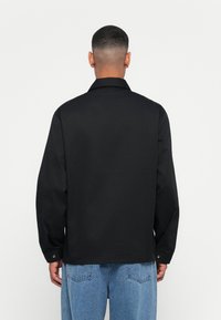 Svart, langermet jakke med krage, knappede mansjetter og en avslappet passform. Glatt tekstur med et ensfarget design, båret over lyseblå jeans.