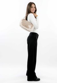 Beige kabelka s umělou kožešinou, lesklým povrchem a horním uchy. Model pózuje s kabelkou přes rameno, v černých kalhotách a bílém topu.