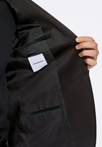 Blazer nero con fodera in satin, caratterizzato da un'etichetta bianca e una tasca di accento verde scuro. Design slim fit con texture liscia.