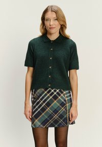 Dunkelgrüner, kurzärmeliger Cardigan mit Kragen, Knopfverschluss und geripptem Saum. Getragen über einem karierter Minirock in Blau, Grün und Braun.