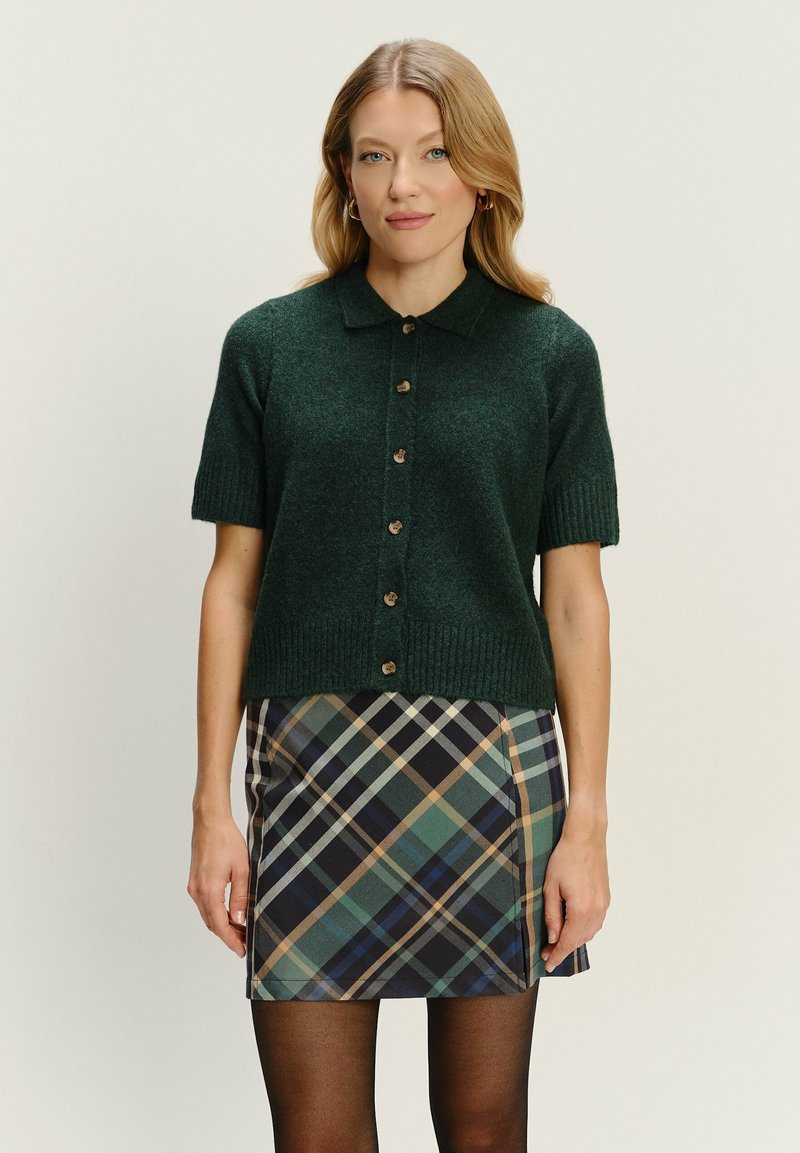 Dunkelgrüner, kurzärmeliger Cardigan mit Kragen, Knopfverschluss und geripptem Saum. Getragen über einem karierter Minirock in Blau, Grün und Braun.