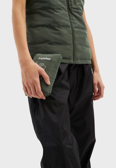 Grön pufferväst med hög krage, dragkedjestängning; hand som håller en kompakt, liknande grön väska med vit grafik. Svarta byxor syns.