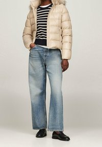 Beżowa kurtka puchowa z kapturem obszytym futrem, granatowo-biały sweter w paski, jasnoniebieskie jeansy o szerokich nogawkach oraz czarne mokasyny.