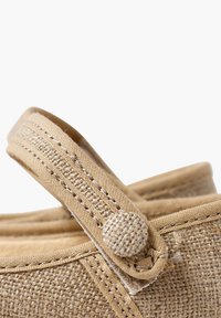 Scarpa in tessuto beige con una tracolla a cerchio, superficie testurizzata e dettaglio a pulsante. Materiale morbido e forma arrotondata.