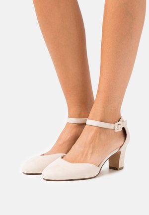 Pieds portant des talons beiges à bout fermé avec bride à la cheville et talons épais sur fond blanc.