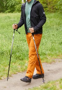 Escursionista con giacca nera e shirt verde, che indossa pantaloni arancioni e scarpe da trekking nere, che utilizza due bastoni da trekking neri su un sentiero di terra.