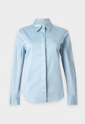 Kvinde iført en lyseblå skjorte med knapper, vidde mørkeblå jeans og hvide spidse sko, poserer med den ene hånd i lommen.