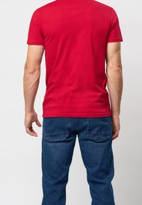 Man wearing a plain red short-sleeve t-shirt and blue denim jeans, standing with his back facing the camera against a white background.