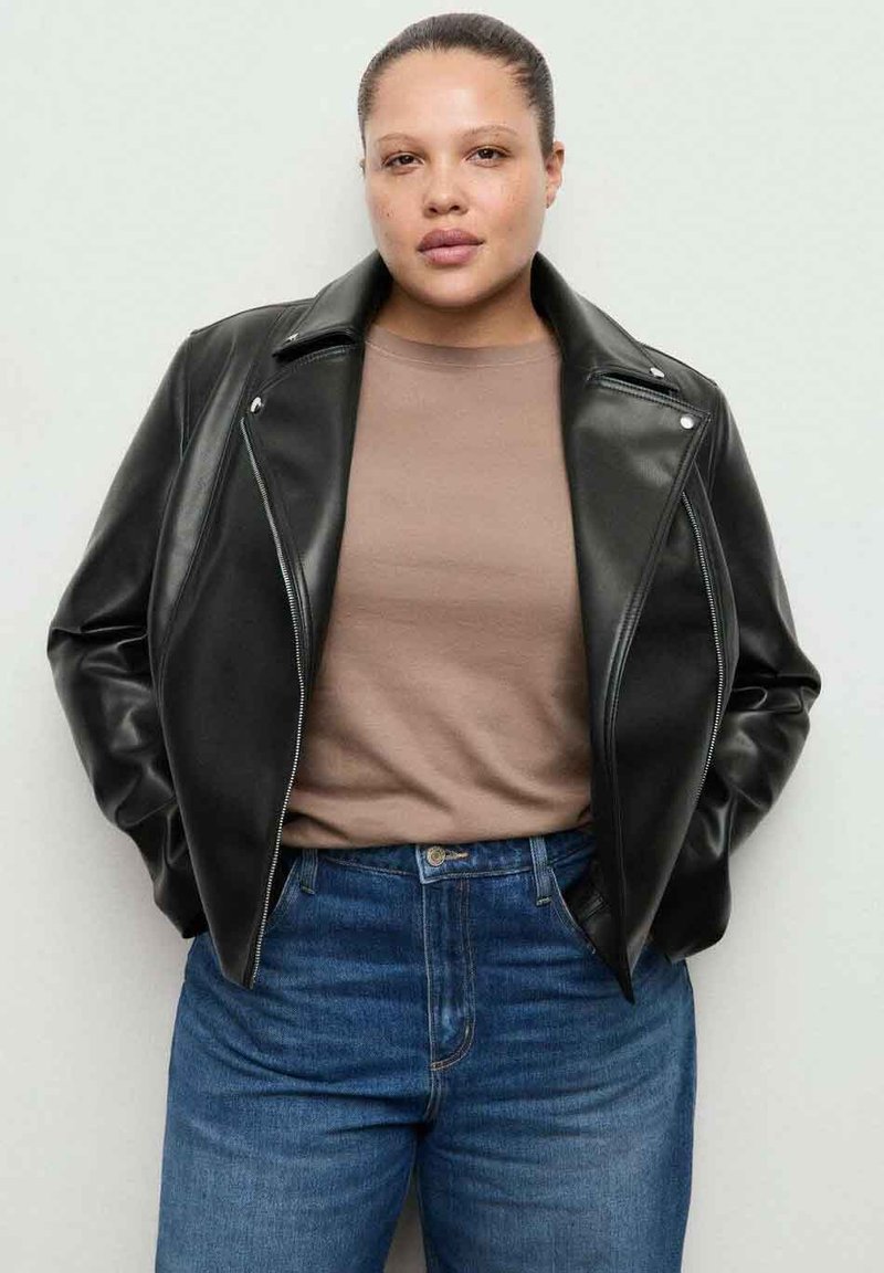 Czarna skórzana kurtka motocyklowa z wyciętym kołnierzem i zapinana na zamek, noszona na beżowej koszuli i niebieskich, wysokich dżinsach.