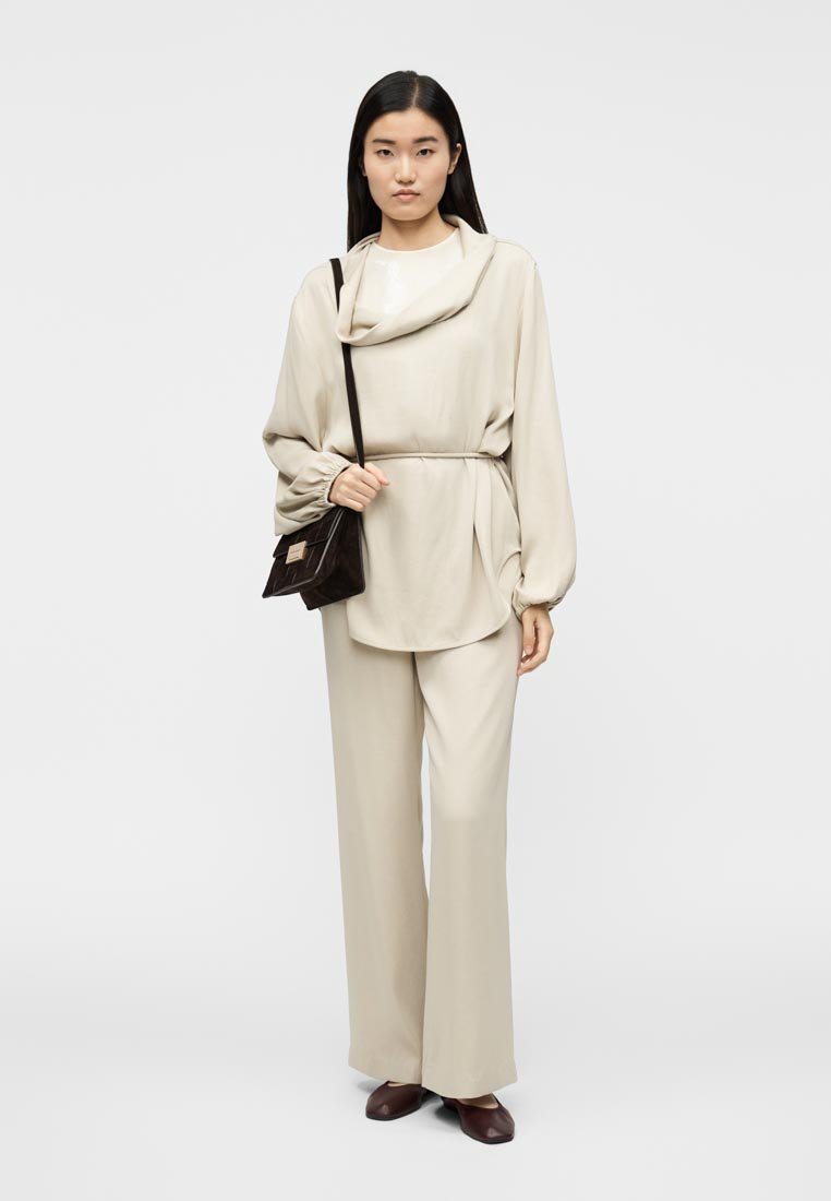 Haut beige fluide à col bénitier et manches froncées, associé à un pantalon ample assorti. Sac à main foncé et chaussures marron complètent le look.