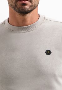 Hellgrauer Pullover mit geripptem Rundhalsausschnitt und einem sechseckigen schwarz-gelben Aufnäher auf der linken Brust. Glatter, strukturierter Stoff.