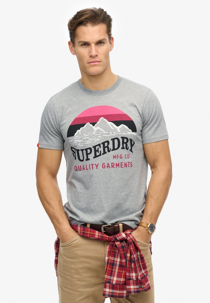 Gray cotton t-shirt with mountain graphic and "SUPERDRY" text in black and red. Model wears beige pants and a red checkered shirt tied around waist.