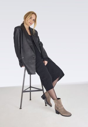 Black leather double-breasted blazer over a black textured dress, paired with brown suede ankle boots featuring a button detail and block heel.