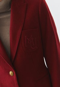 Punane villane bleiser, millel on nurgakolmnurkne krae, kuldne nupudetail ja tikitud tasku, millel on "MU" initsiaalid. Sile tekstuur, kohandatud disain.