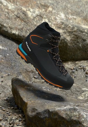 Fekete és narancssárga túracipő kék díszítéssel, fűzőkkel és masszív talppal, sziklás terep felett lebegve.