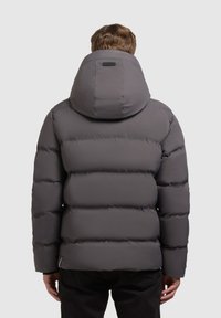 Grijze pufferjack met een dikke, gewatteerde ontwerp en een grote capuchon. Kenmerken zijn een zwart merkembleem op de achterkant en elastische manchetten.