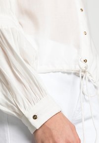 Blusa branca em tecido leve e transparente, com mangas gather e punhos com botões dourados. Cordão ajustável na bainha.