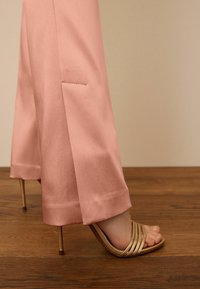 Pantalons en satin rose clair avec une fente sur le côté, associés à des sandales à talons hauts dorées à lanières. Texture lisse, design élégant, détails minimalistes.