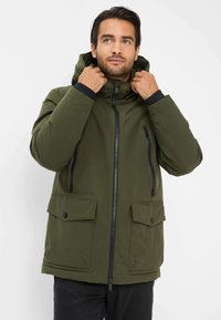 Giacca isolante verde con cappuccio, chiusura zip frontale, due ampie tasche frontali e dettagli neri sulle cerniere e sui polsini. Texture liscia.