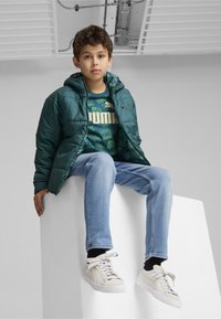 Chaqueta acolchada verde con capucha, combinada con una camiseta gráfica verde oscuro y vaqueros azul claro. Zapatillas blancas completan el conjunto.