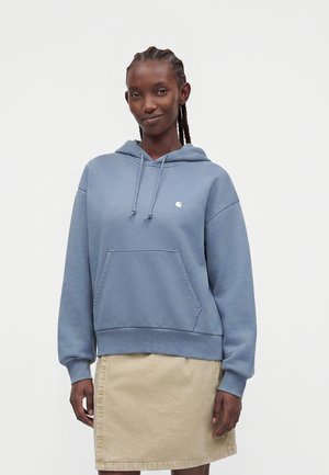 Femme aux cheveux tressés portant un sweat à capuche bleu et une jupe beige, debout devant un fond clair uni.