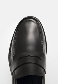 Mocassin en cuir noir poli avec un bout cousu et une bride décorative traversant le dessus, sur fond blanc.