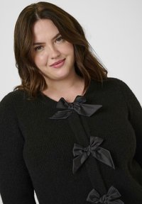 Schwarze gestrickte Strickjacke mit drei Satin-Schleifenakzenten auf der Vorderseite. Weicher Stoff mit einem gerippten Muster und einem Rundhalsausschnitt.
