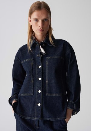 Dark blue denim jacket with a button-down front, two chest pockets, and contrasting stitching. Features a collar and relaxed fit.